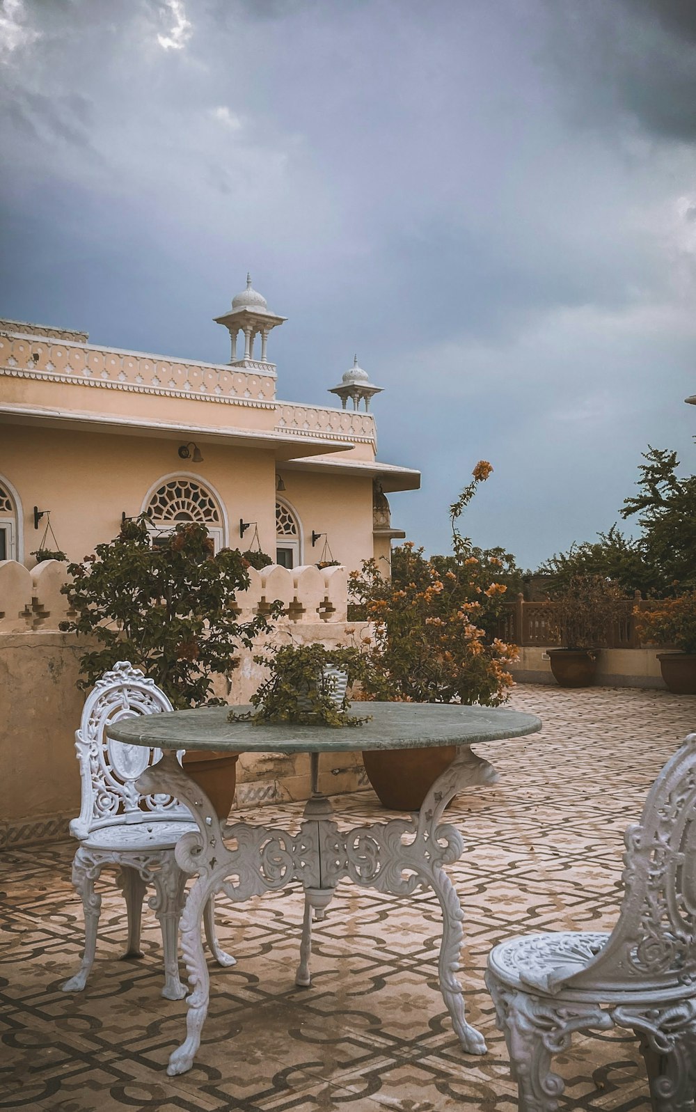 Wrought-iron chairs around a stone table on the tiled rooftop terrace of a haveli, under a gathering monsoon sky
