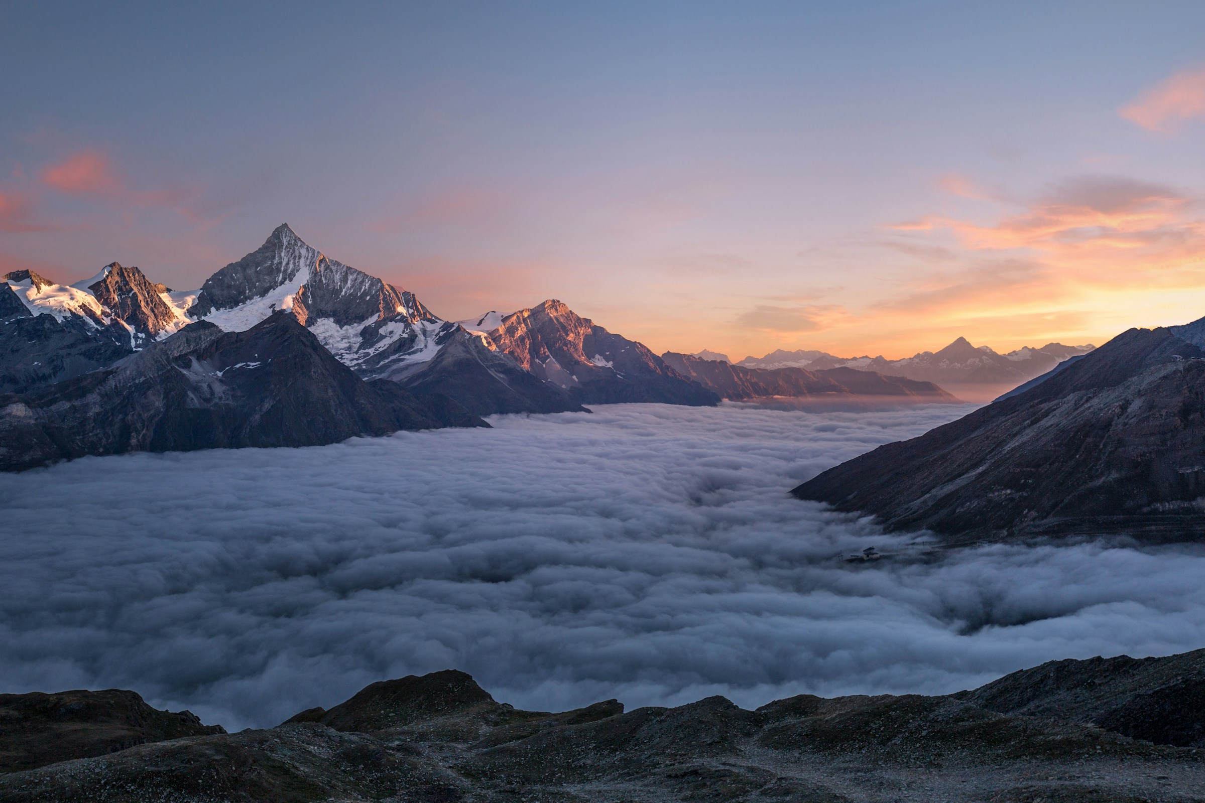 Snow peaks catching pink-gold alpenglow above a valley filled with cloud.